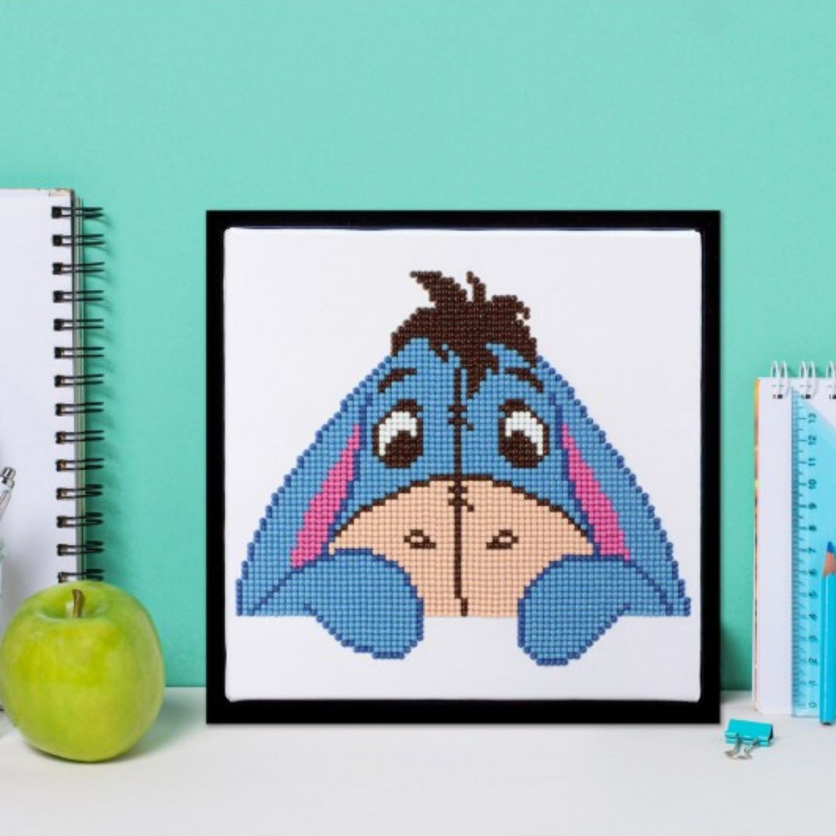 Completed Eeyore diamond painting displayed in a black frame sitting on a desk next to a green apple, notebooks, and pens.