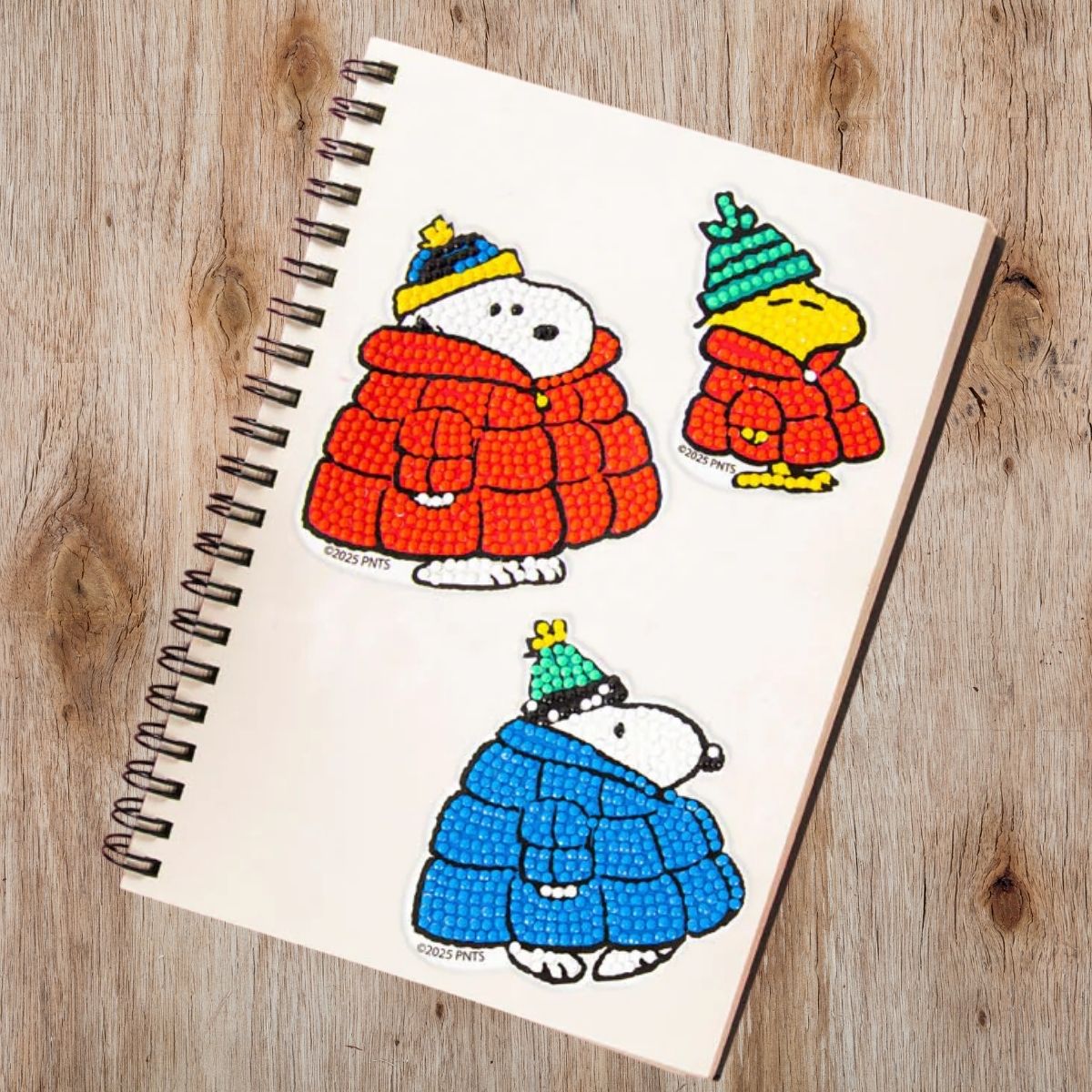 Three completed Peanuts winter diamond painting stickers applied to the cover of a blank spiral notebook resting on a wooden desk.
