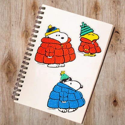 Three completed Peanuts winter diamond painting stickers applied to the cover of a blank spiral notebook resting on a wooden desk.