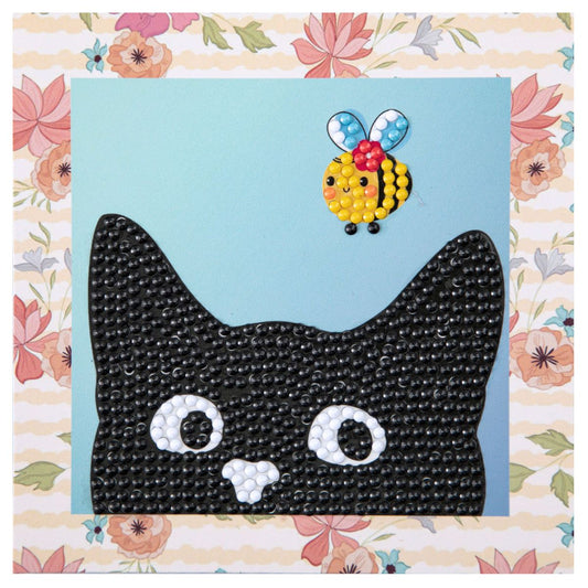 Finished DIAMOND DOTZ Mischievous Black Cat diamond painting featuring a black cat peeking at a bumblebee with a peach floral border.