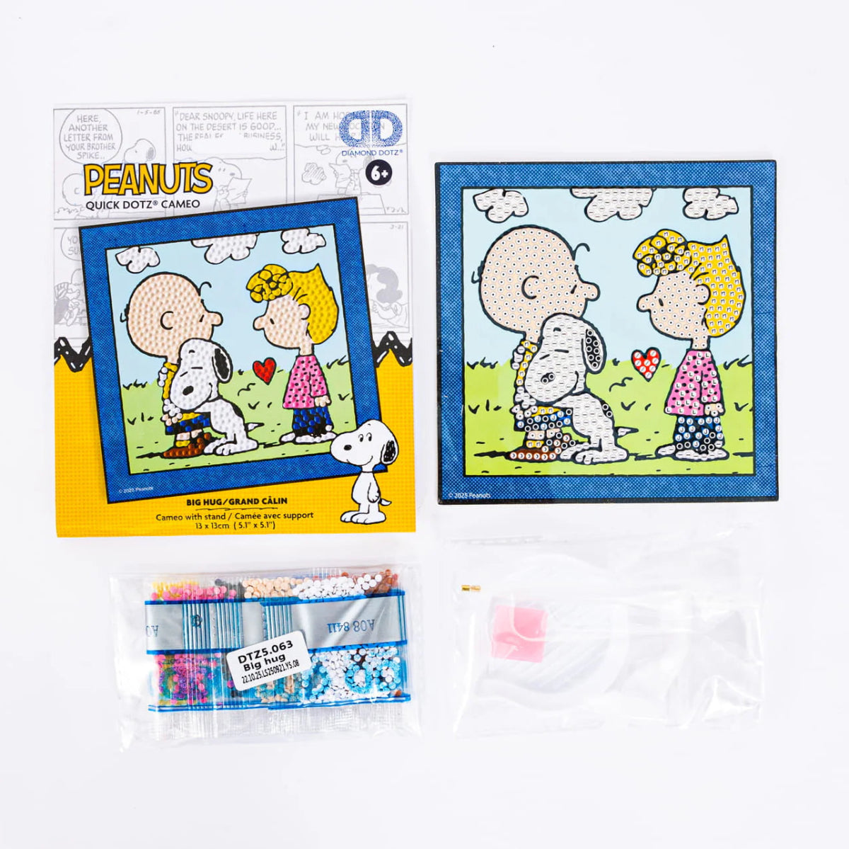 Retail packaging and contents of the Diamond Dotz Peanuts Big Hug kit, including the canvas, colorful diamond drills, stylus tool, wax, and craft tray.