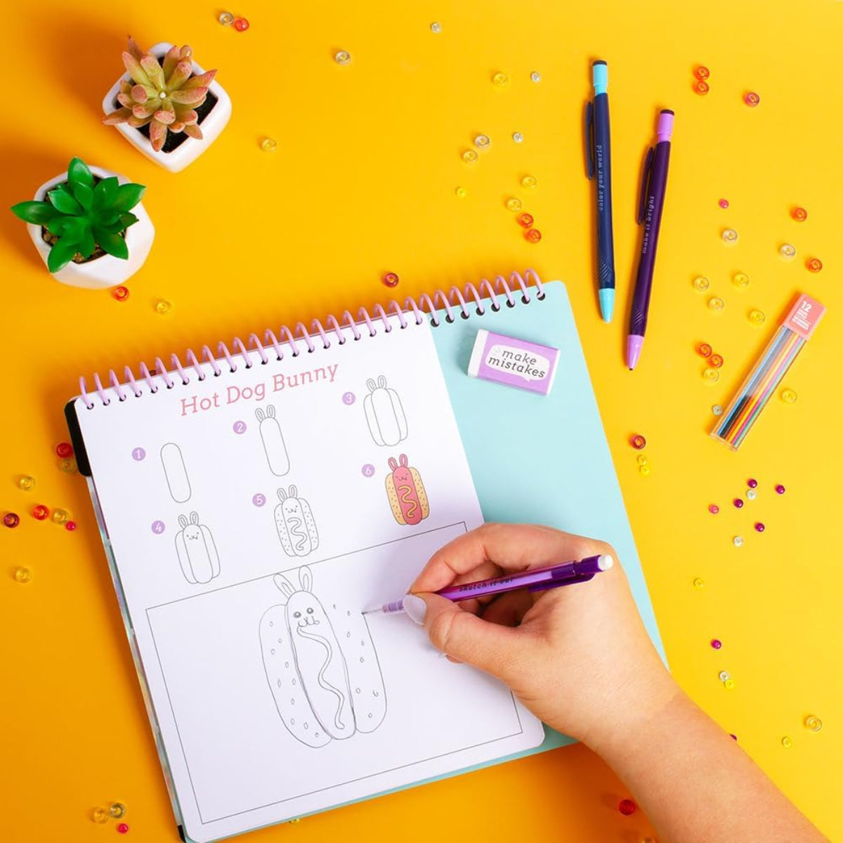 Person coloring a 'Hot Dog Bunny' design in a spiral-bound notebook with colorful pens on a yellow background.