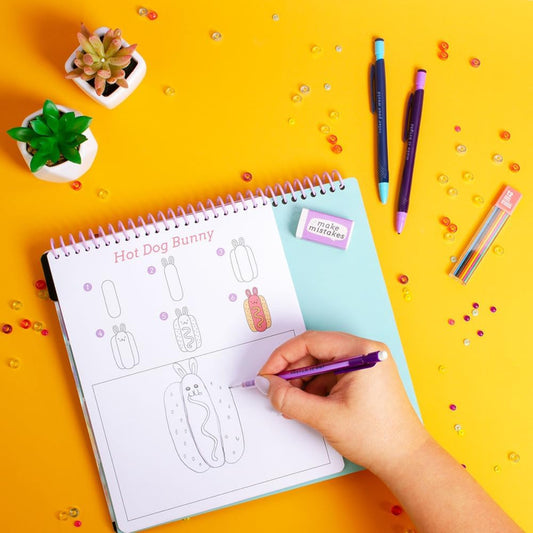 Person coloring a 'Hot Dog Bunny' design in a spiral-bound notebook with colorful pens on a yellow background.