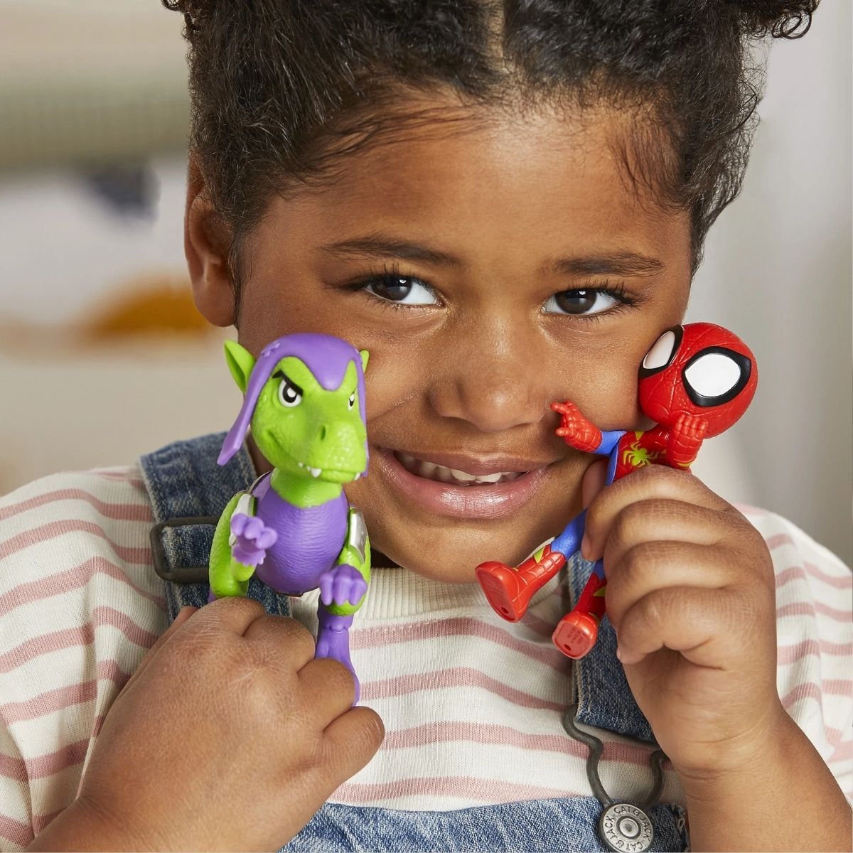 Close-up lifestyle shot of a smiling young child playfully holding the Spidey figure and Goblin Raptor toy near their face.