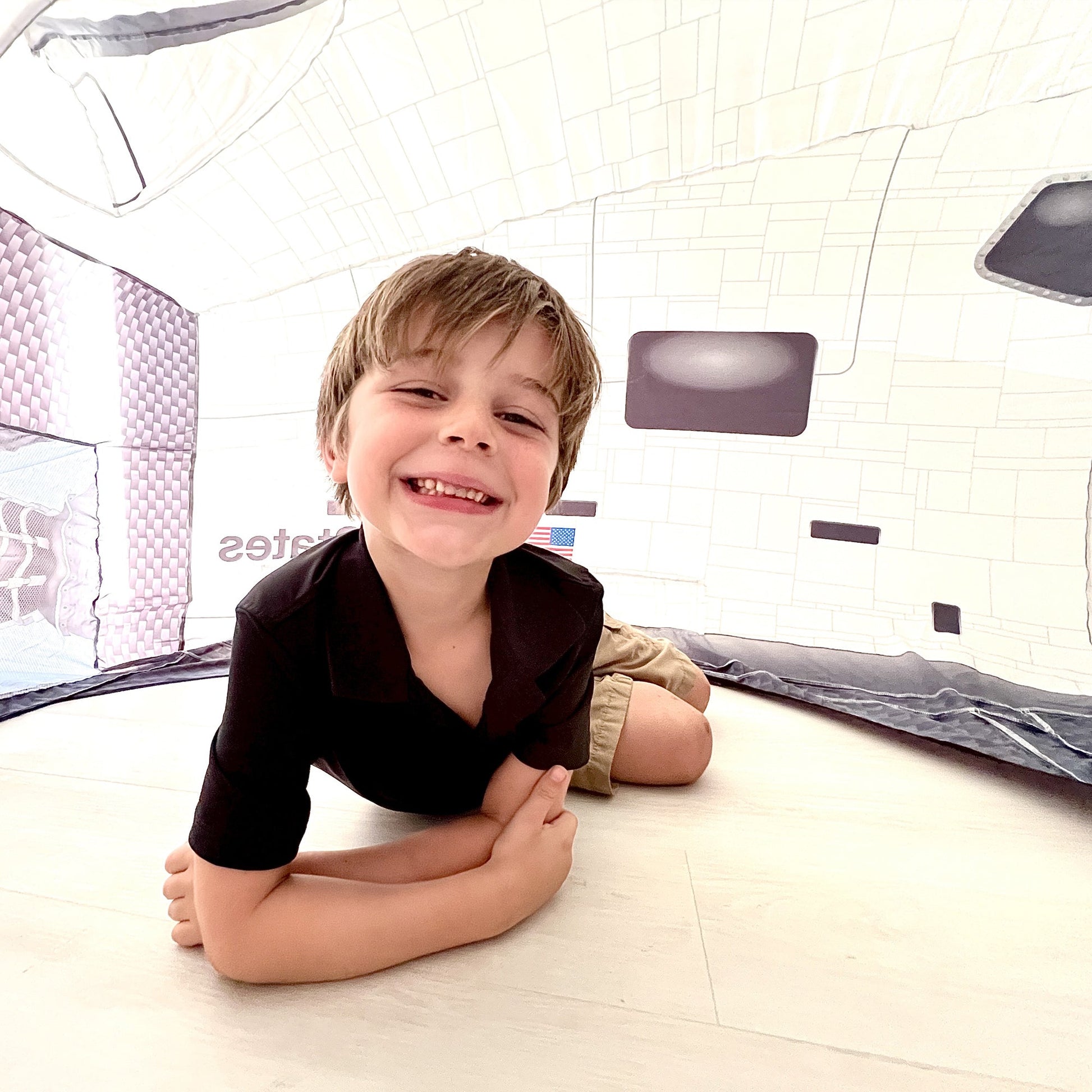 A young boy lying on his stomach inside the Space Shuttle AirFort smiling widely at the camera.