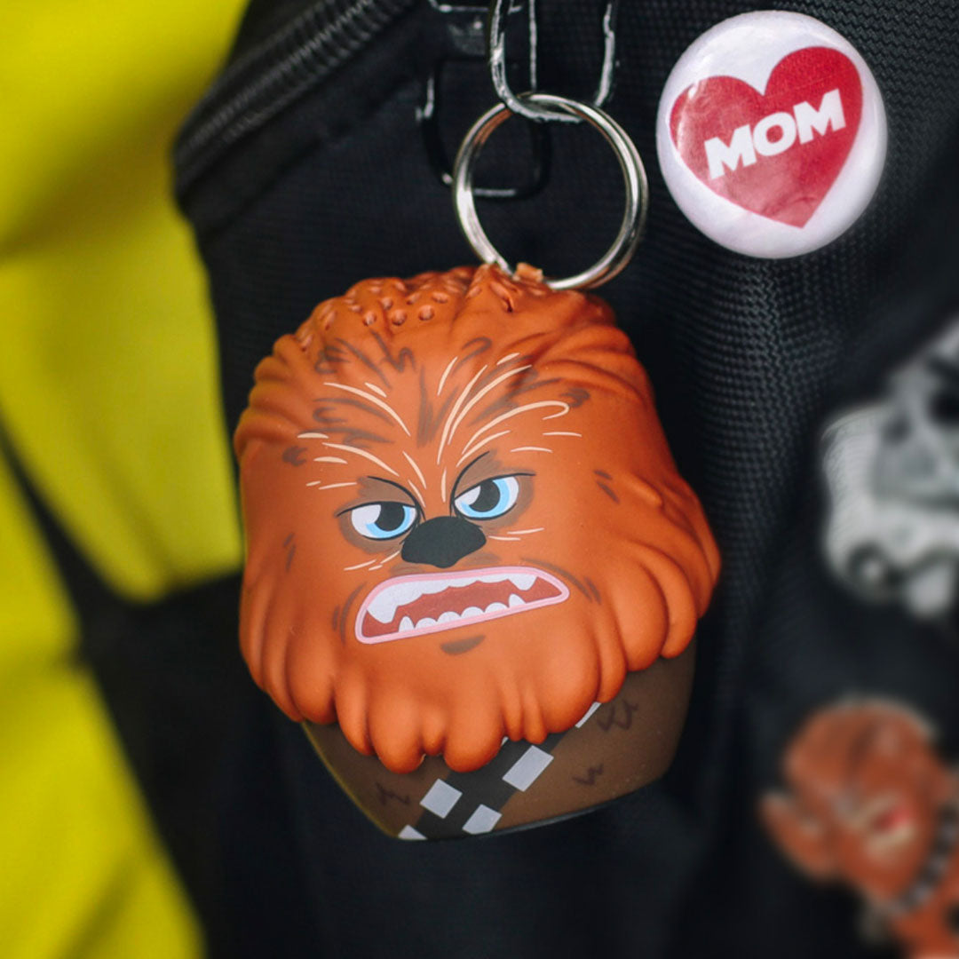 Lifestyle shot of the Chewbacca mini Bluetooth speaker attached to a black backpack using a keychain ring, sitting next to a 'MOM' pin.