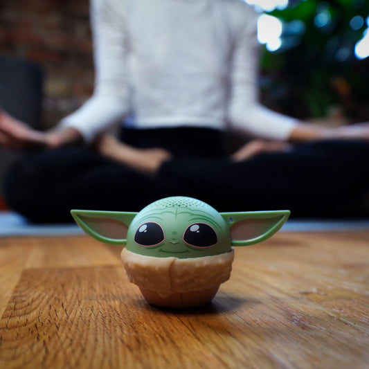 Lifestyle shot of the Grogu mini Bluetooth speaker sitting on a wooden floor with a person meditating in the blurred background.