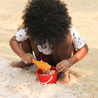 Child crouching in the sand using the yellow scoop and red bucket.