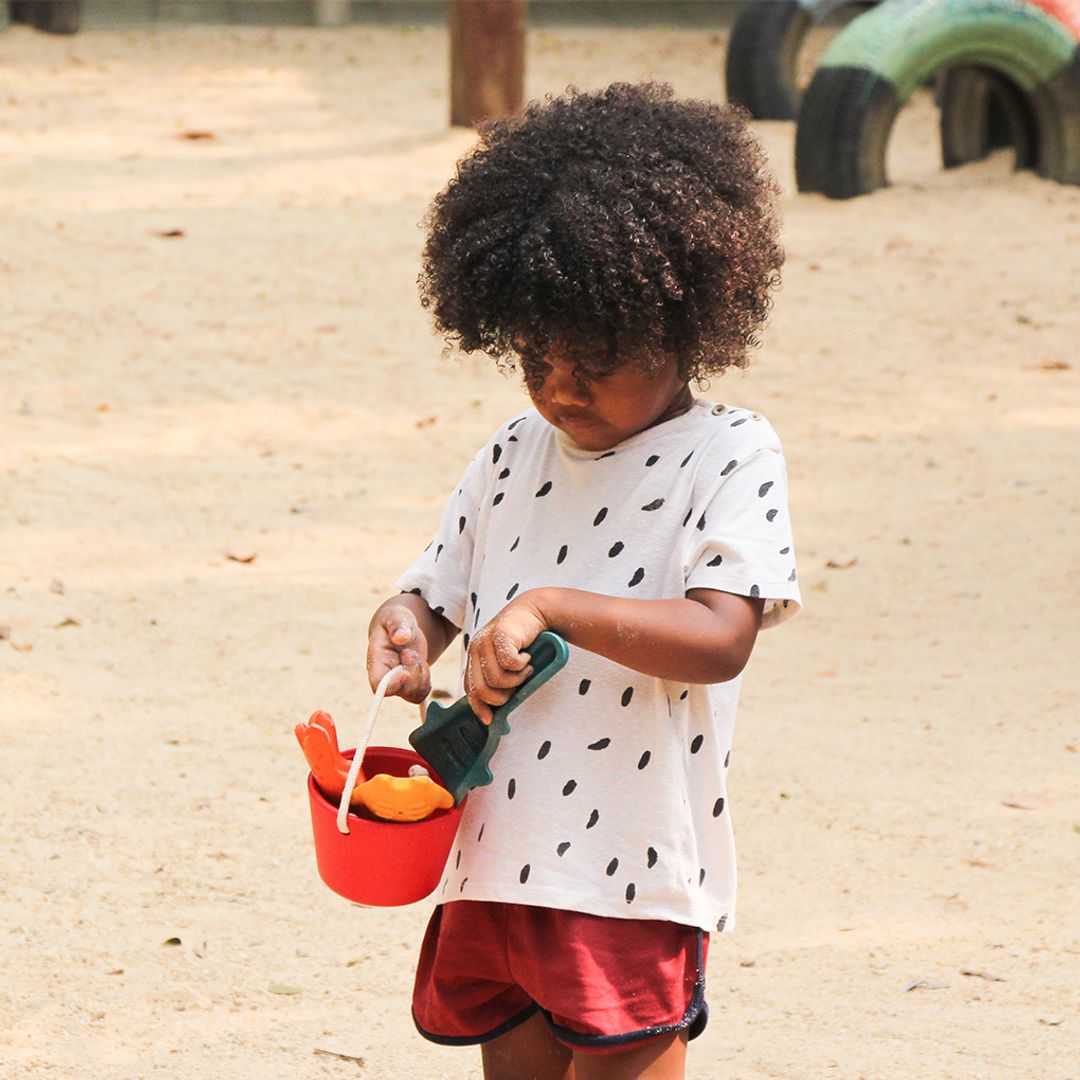 Child standing outdoors holding the red bucket and colorful wooden sand tools.