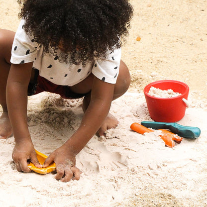 Child sitting in beach sand playing with the PlanToys wooden sand tools and bucket.