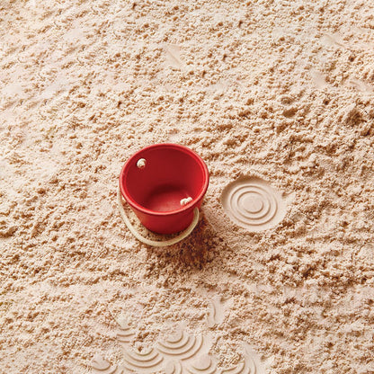 Red PlanToys wooden sand bucket resting on sand next to a circular bucket imprint.