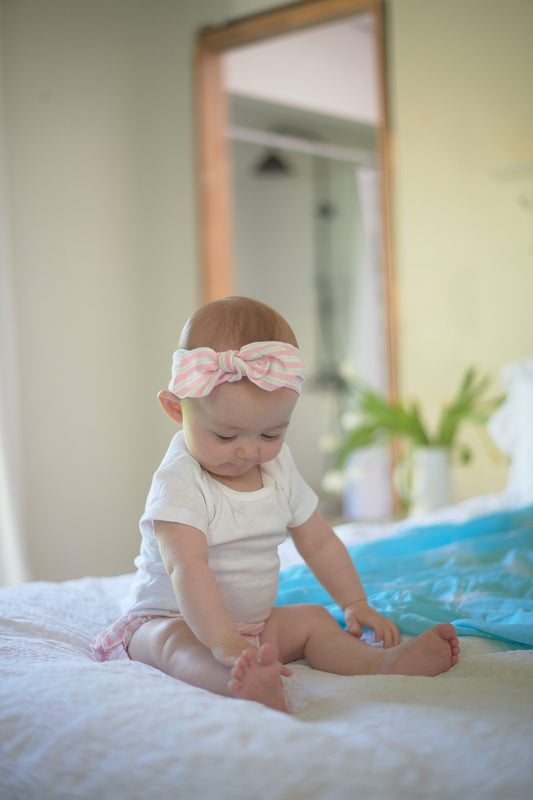 Baby girl wearing the Newcastle Classics Candy Stripe headband and white onesie sitting on a bed.